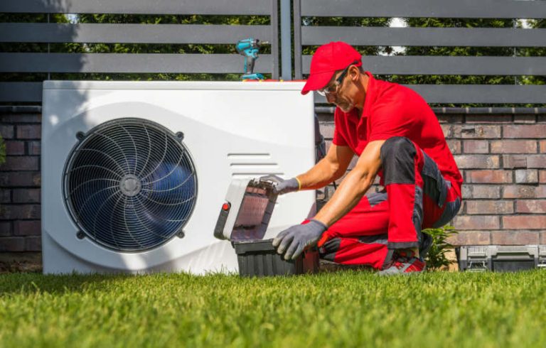 A technician in a red uniform and cap kneels on a green lawn to open a toolbox next to an outdoor air conditioning condenser unit.