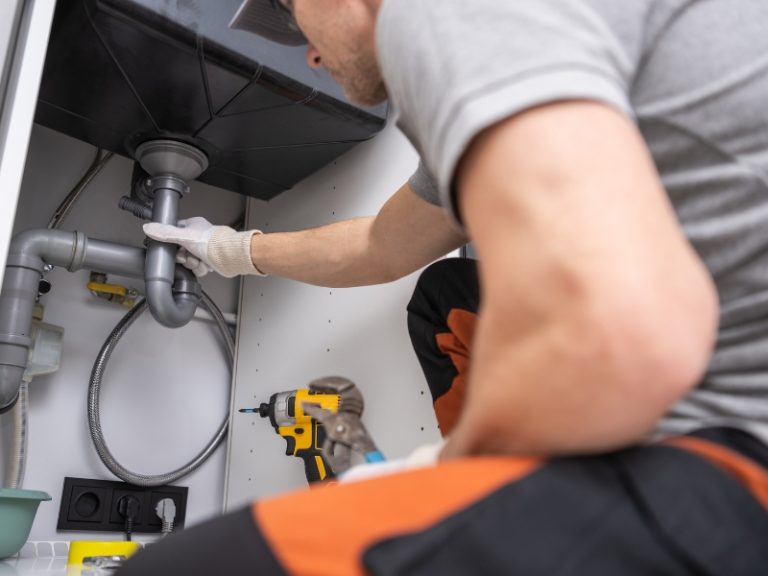 A plumber or technician wearing gloves and a grey uniform working under a kitchen sink. He is holding a grey PVC P-trap pipe, with an impact driver visible in the foreground. The focus is on the sink's drainage assembly within a white cabinet.